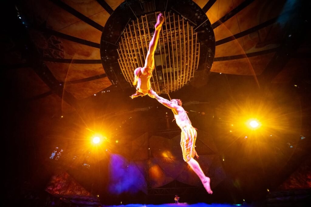 Two performers engage in an aerial hoop act, silhouetted by dramatic stage lighting and warm orange spotlights.