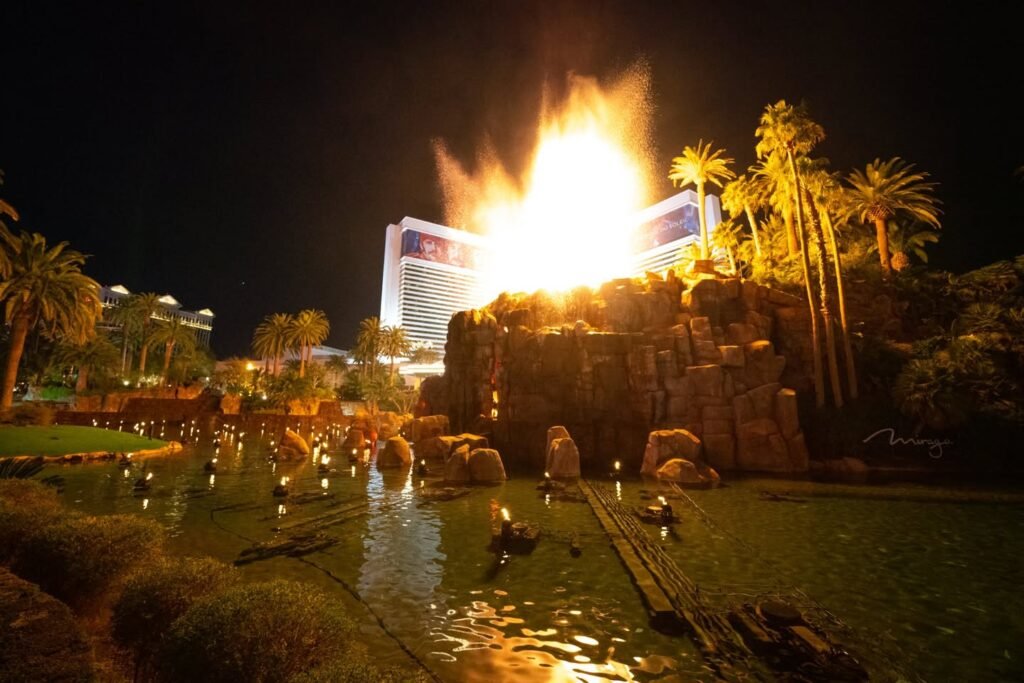 A nighttime view of a resort with a large fire display above palm trees and a reflecting pool.