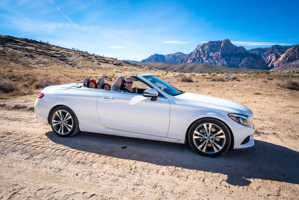 A white Mercedes-Benz convertible driving on a sandy road in a desert with mountains in the background under a clear blue sky.