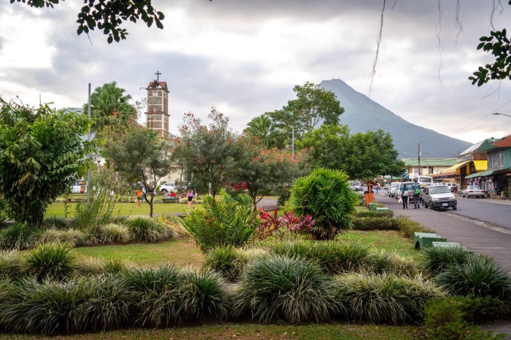 A view of a maintained public park in front of a street with a church tower and mountain in the distant background under cloudy skies.
