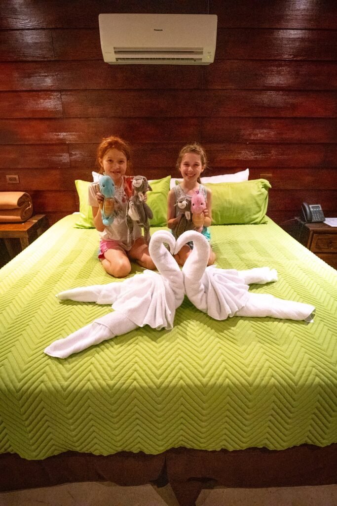 A young child sits on a hotel bed with a towel folded into a heart-shaped swan figures.