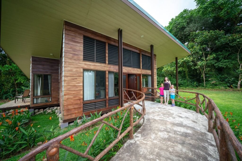 A modern house with wooden walls and a green roof stands in a lush tropical garden, accessed by a charming wooden walkway.