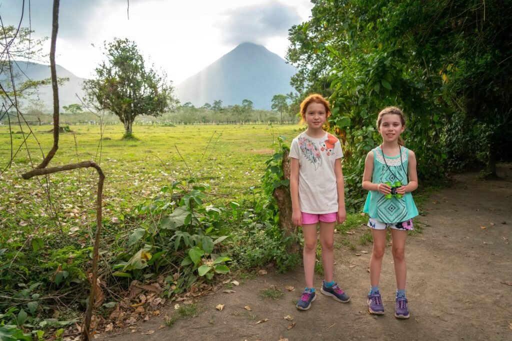 Two hikers stand on a dirt path near lush green walls and distant volcanic peaks.