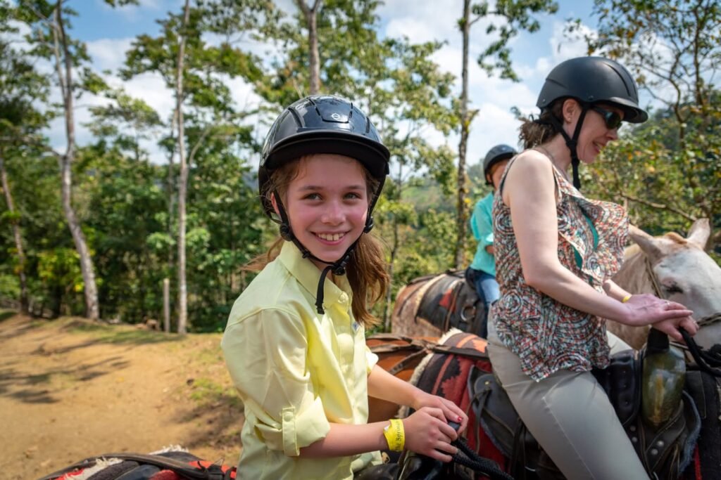 People wearing helmets ride horses along a trail surrounded by tall trees and greenery under a bright sky.