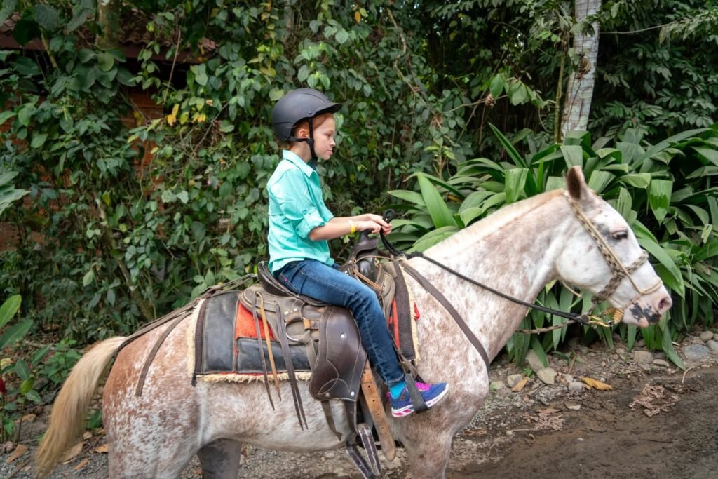 A young rider on a dappled gray horse in a lush forest setting, wearing a helmet and jeans while enjoying a scenic trail ride.