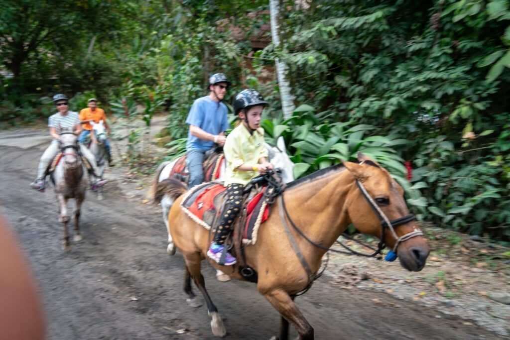 Riders on horseback travel along a dirt path through lush green foliage in a tropical setting.