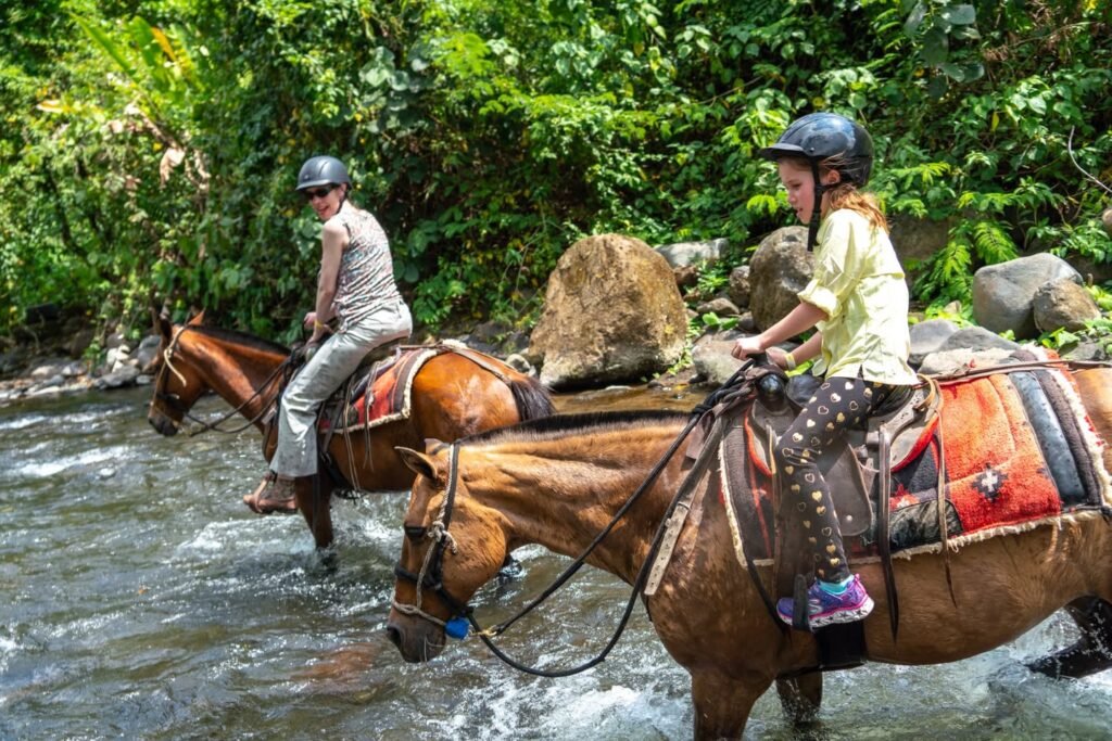 Two riders in safety helmets guiding horses through a shallow, rocky stream.