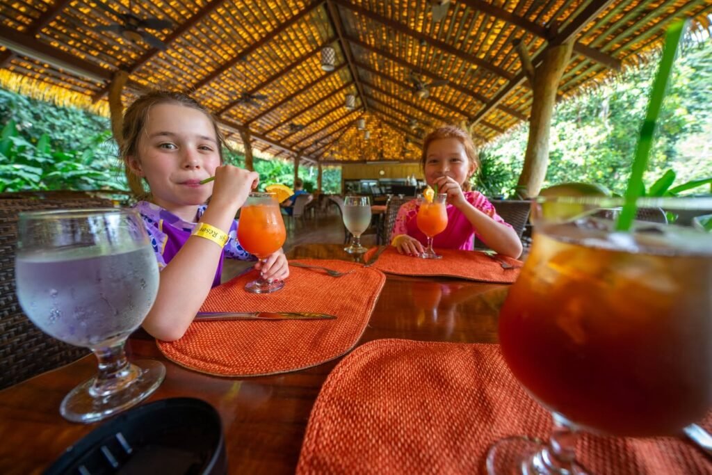 Two children enjoy frozen cocktails at a jungle restaurant with a thatched roof.