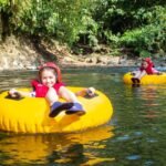 Horseback Riding at Club Rio Two people wearing red helmets tubing down a calm river on bright yellow inner tubes.