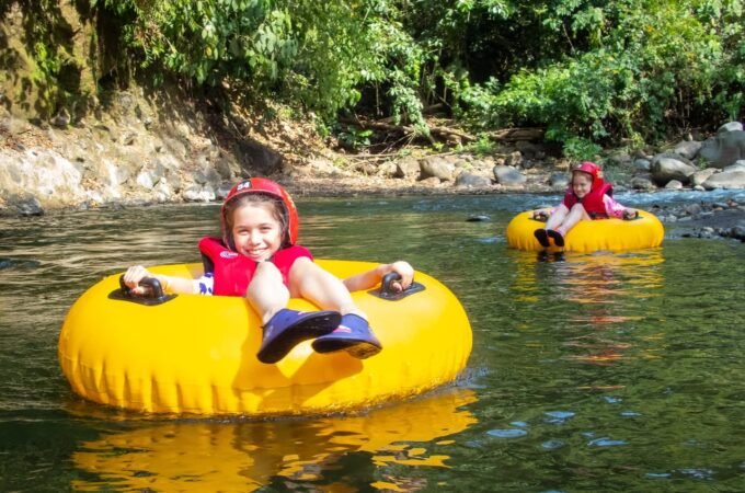 Two people wearing red helmets tubing down a calm river on bright yellow inner tubes.