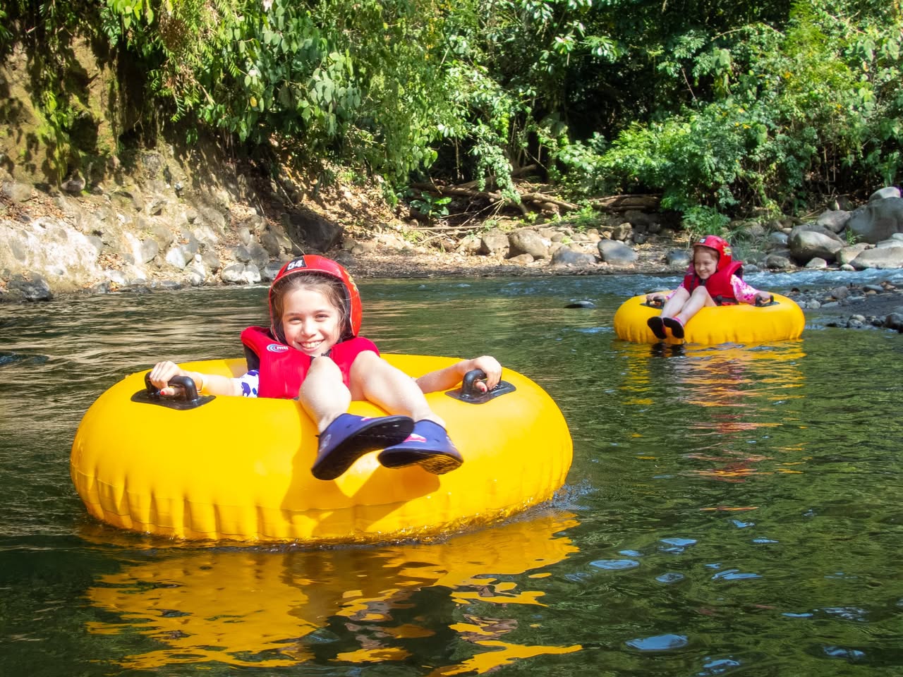 Two people wearing red helmets tubing down a calm river on bright yellow inner tubes.