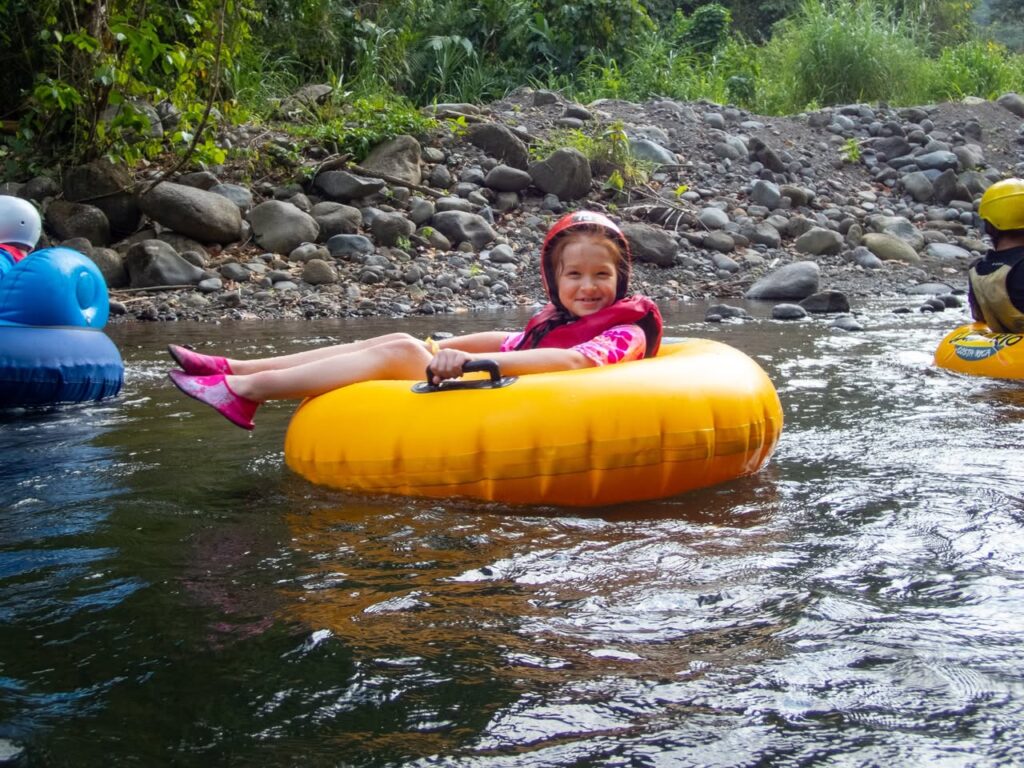 A child wearing a helmet and life jacket floats on a yellow inner tube in a river.