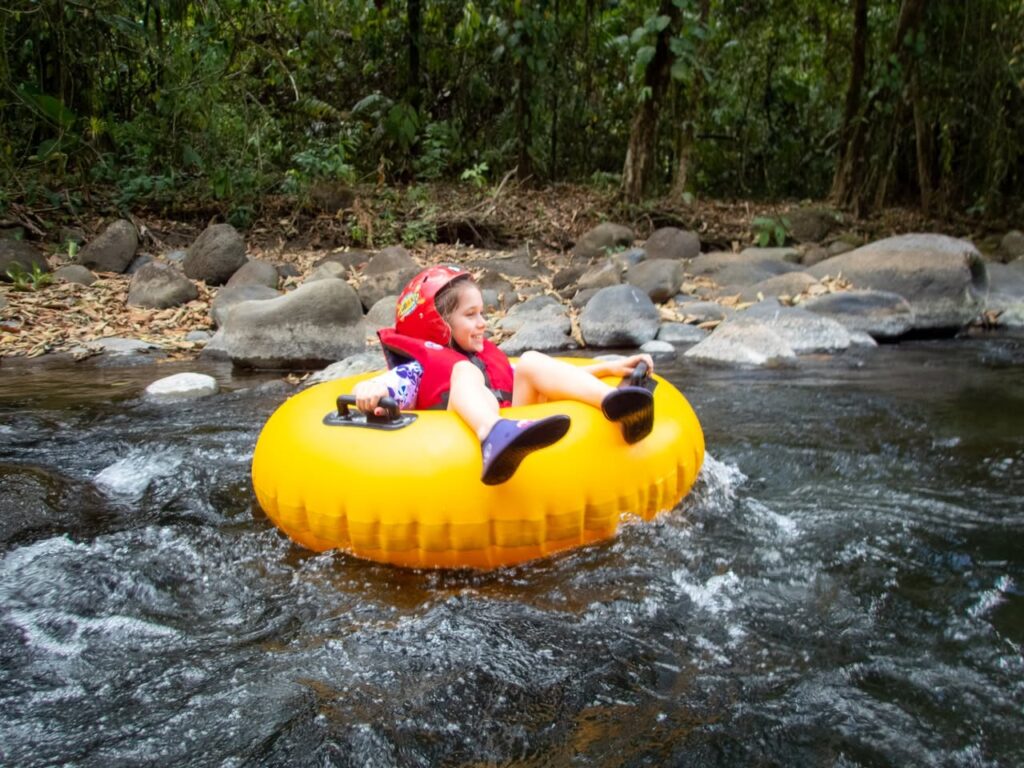 A person in a red life jacket rides a bright yellow tube down a flowing stream. The riverbank is lined with rocks and lush green vegetation.