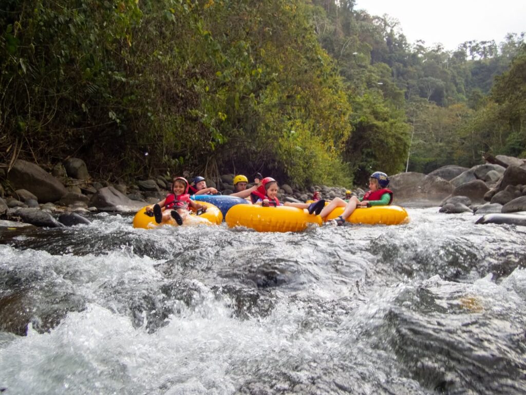 People navigate a rocky river in colorful inner tubes. The scene shows fast-moving rapids surrounded by dense forest.