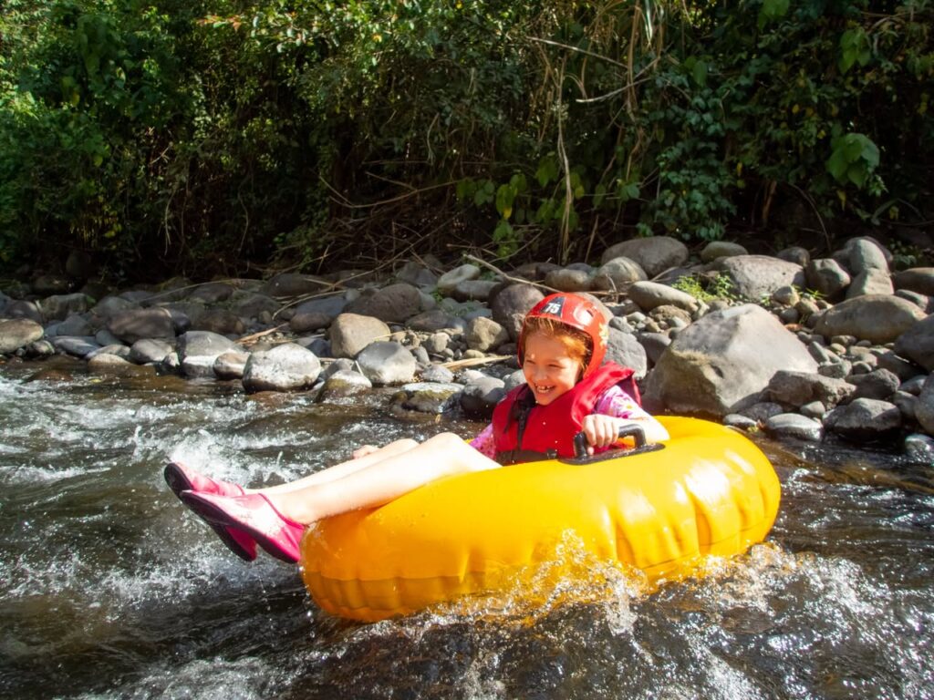 A child wearing a helmet and life jacket smiles while floating down a rocky river in a bright yellow inner tube.
