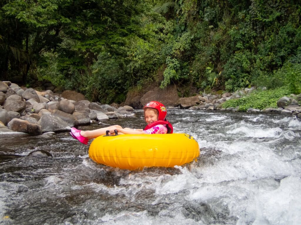 Tubing down a rushing river on a bright yellow inflatable tube, wearing a red helmet, surrounded by greenery and rocks.