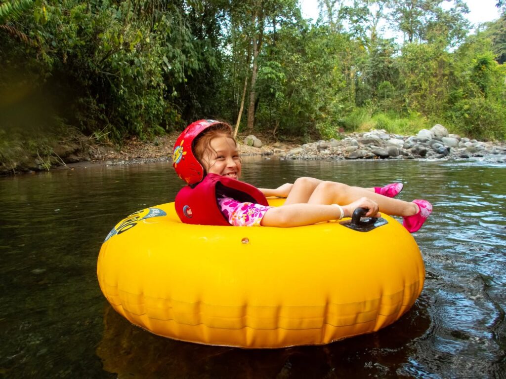 A person floats on a yellow inflatable tube through a calm, tree-lined river. The shoreline features lush green tropical foliage and rocky banks.