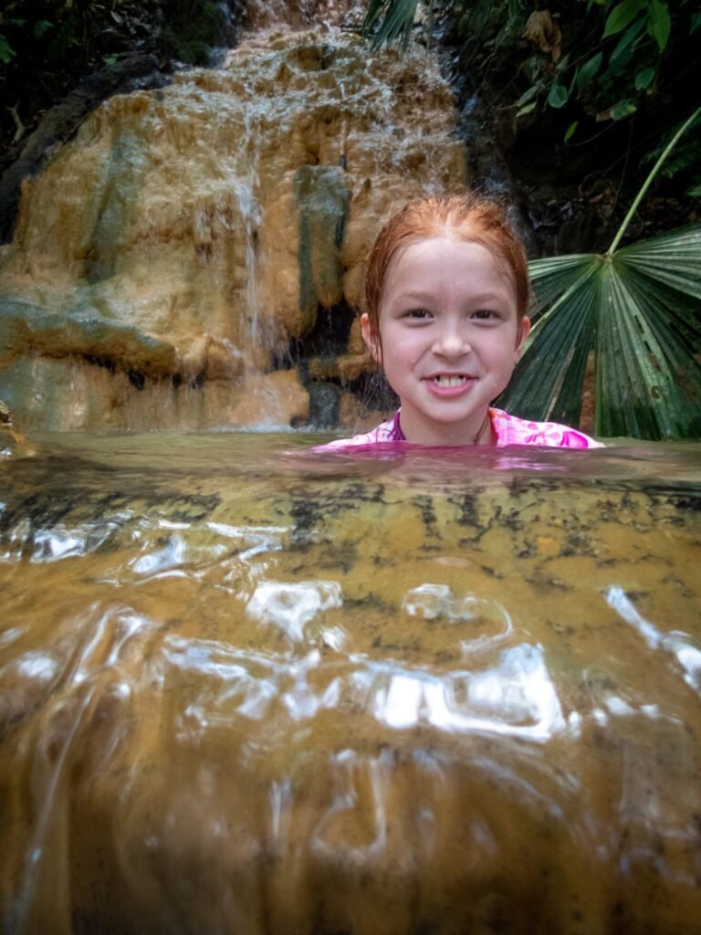 A person soaks in a natural hot spring with a waterfall in the background. Lush tropical foliage surrounds the rocky pool.
