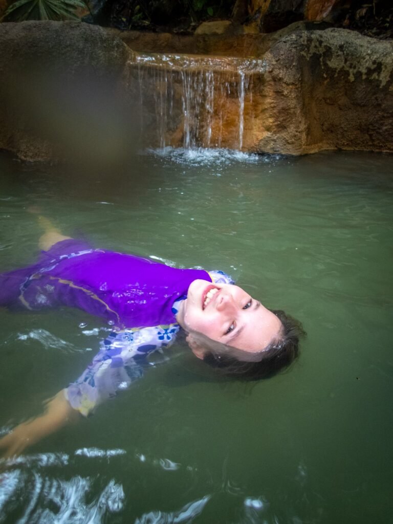 A person floats on their back in a green pool with a small waterfall in the background, wearing a purple swim top.