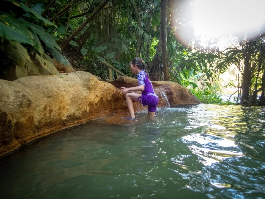 A child in a purple costume stands in a natural green pool surrounded by lush tropical foliage.