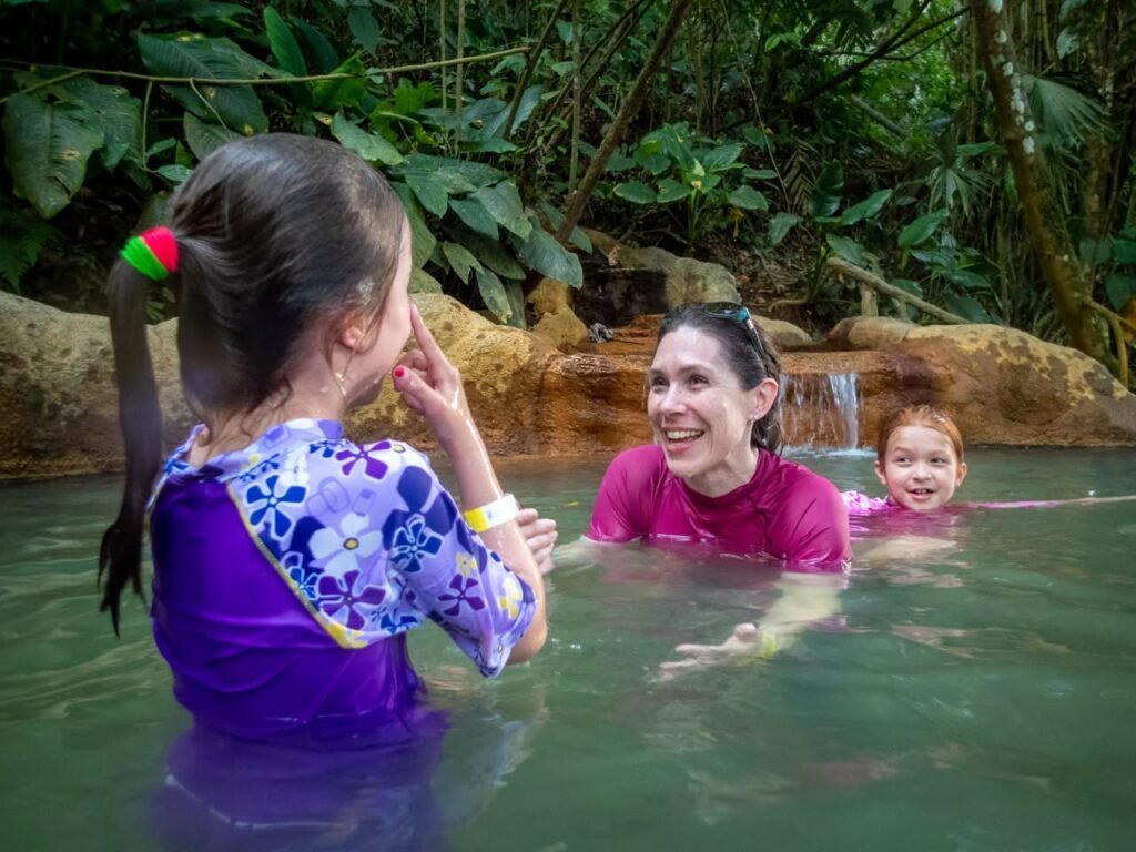 A family enjoys swimming together in a lush tropical thermal pool. Vibrant colors contrast with the natural greenery and stone surroundings.