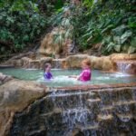 Two people relax in a natural stone pool with cascading waterfalls amidst lush tropical vegetation. The setting features a multi-tiered rock formation and a lush jungle environment.