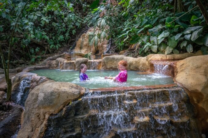 Two people relax in a natural stone pool with cascading waterfalls amidst lush tropical vegetation. The setting features a multi-tiered rock formation and a lush jungle environment.
