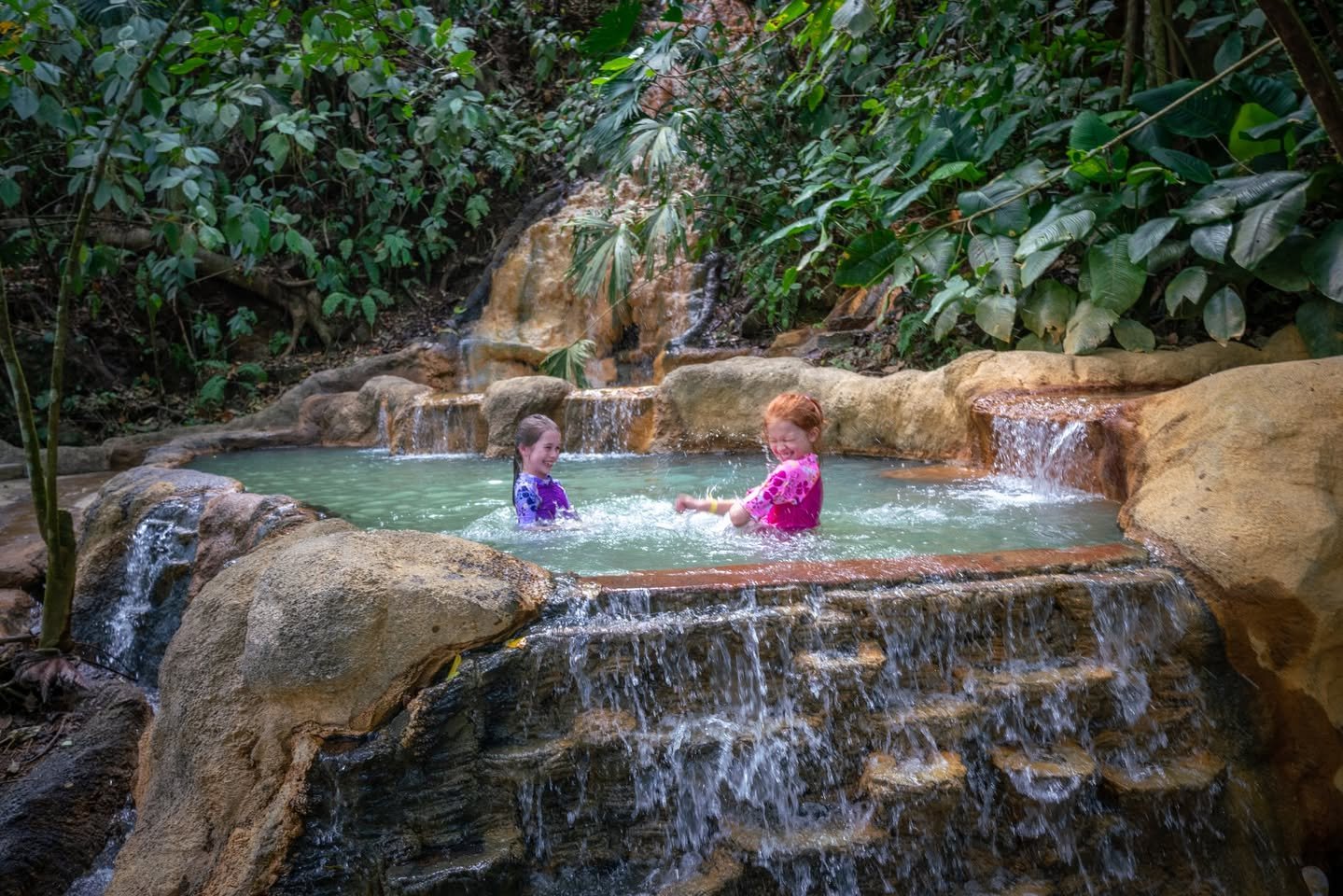 Two people relax in a natural stone pool with cascading waterfalls amidst lush tropical vegetation. The setting features a multi-tiered rock formation and a lush jungle environment.