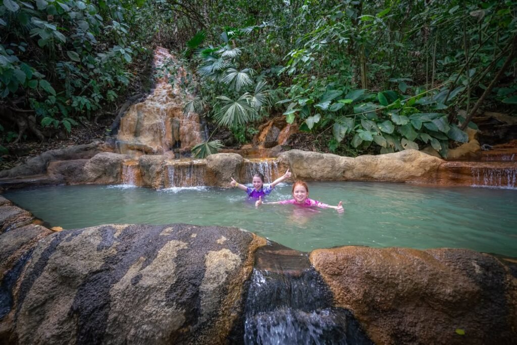 Two people enjoy a natural spring pool surrounded by lush tropical vegetation.