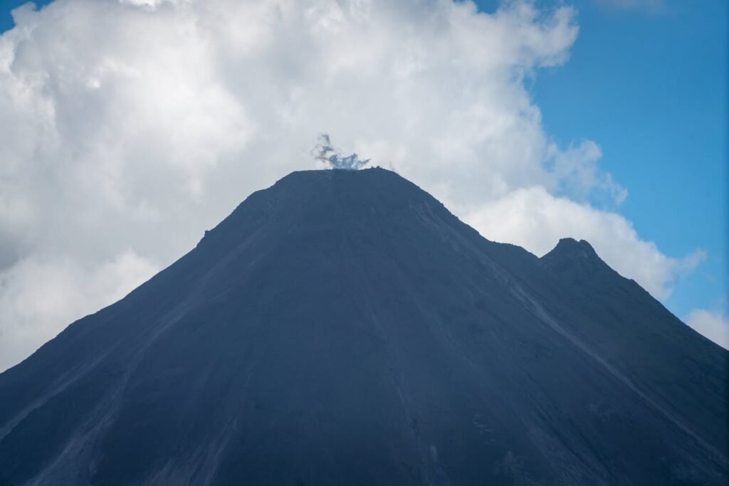 A volcanic peak with plumes of smoke and ash rising from the summit against a partly cloudy sky.