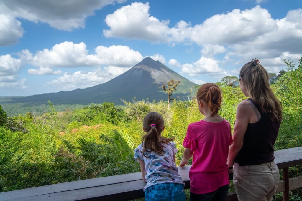 Three people enjoy a view of a mountain peak from a scenic lookout with lush greenery and a bright blue sky.