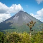 A prominent volcano with a smoking peak rises above a dense green forest under a blue sky. Several white clouds are visible in the sky.