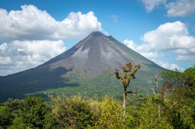 A prominent volcano with a smoking peak rises above a dense green forest under a blue sky. Several white clouds are visible in the sky.