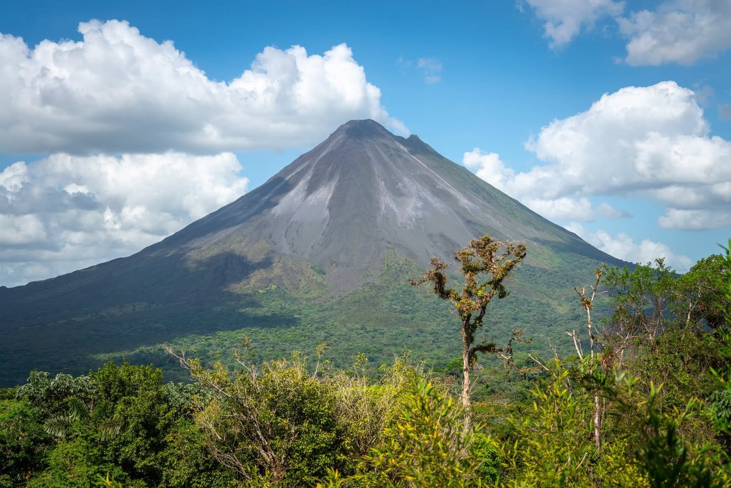 A prominent volcano with a smoking peak rises above a dense green forest under a blue sky. Several white clouds are visible in the sky.