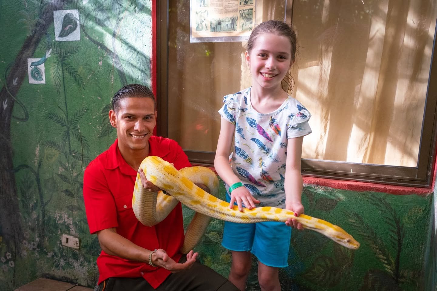 Two individuals holding a large yellow anaconda at a reptile exhibition in Ecuador. The snake has distinct yellow and orange patterns.
