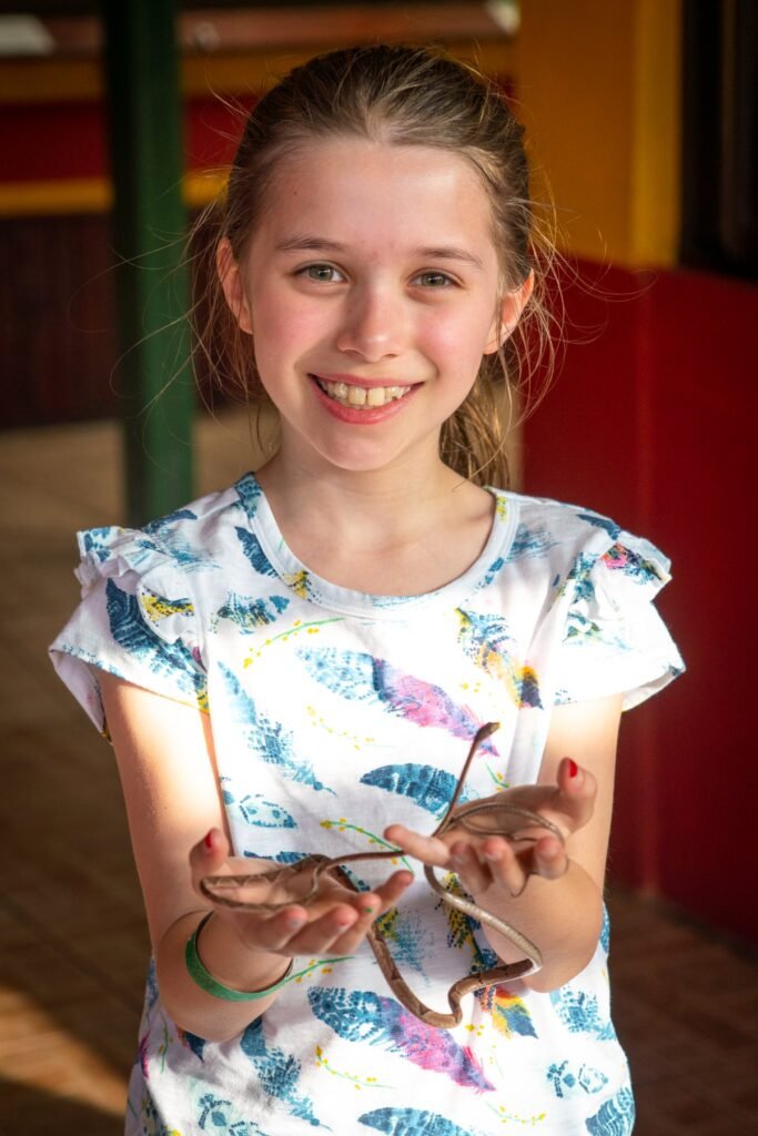 A young person wearing a floral shirt smiles while holding a small lizard and wearing glasses in hand, standing indoors with colorful background elements.