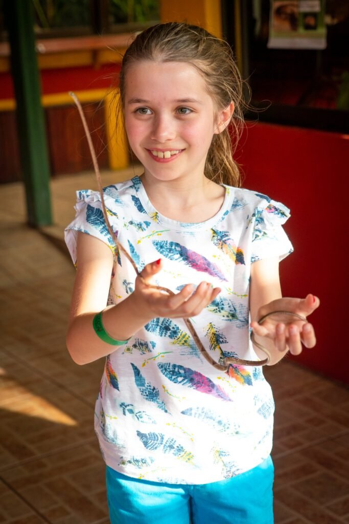Child dressed in a t-shirt with a butterfly motif holding a stick, smiling while standing in an indoor garden setting.