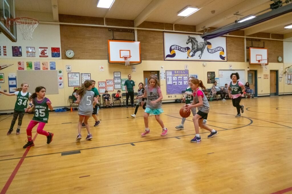 Girls in green and gray jerseys play dynamic basketball in a local school gym during a community game.
