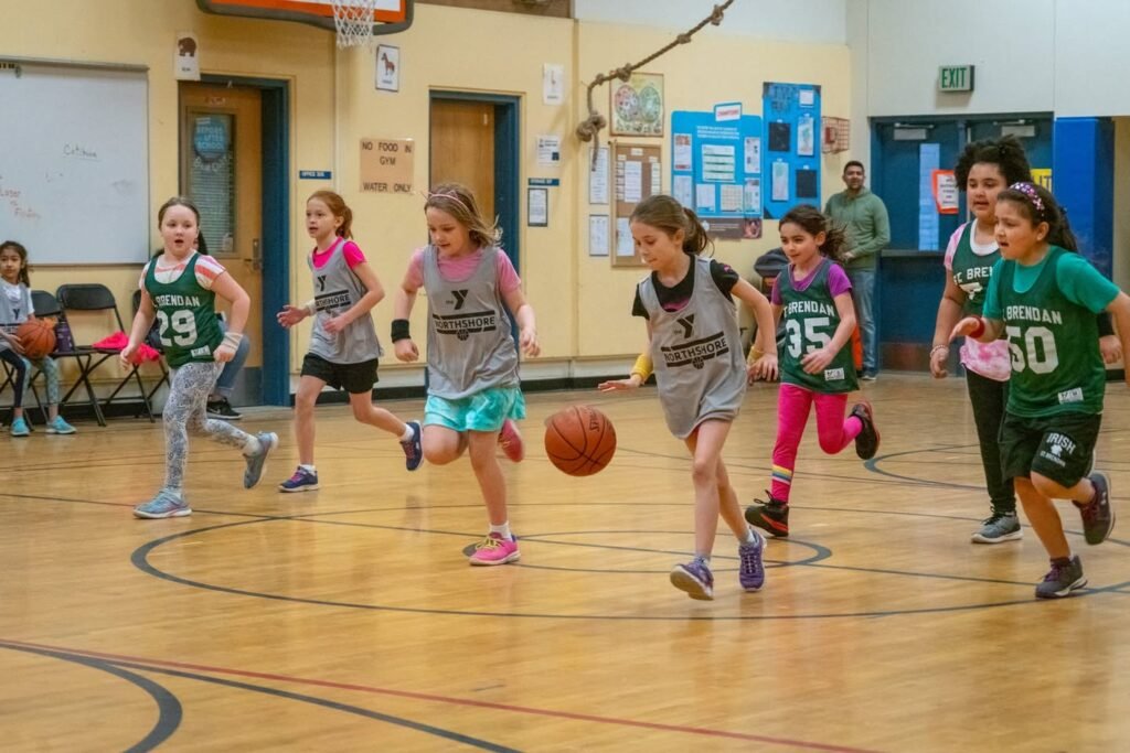 Children playing indoor basketball on a school gym floor with colorful uniforms.