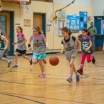 Children playing indoor basketball on a school gym floor with colorful uniforms.