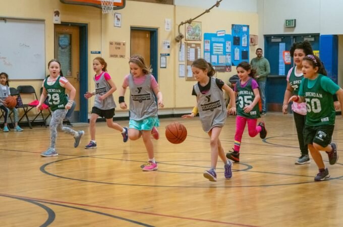 Children playing indoor basketball on a school gym floor with colorful uniforms.