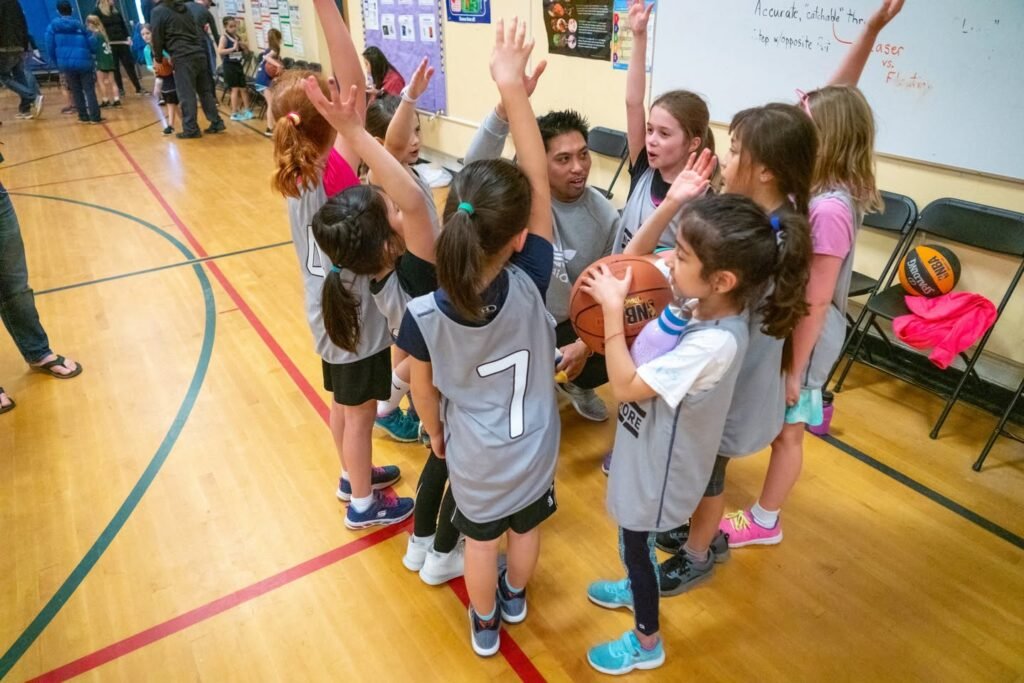 Kids in gray jerseys raise hands in a circle during a basketball practice. A coach holds a basketball while the group focuses on the game.