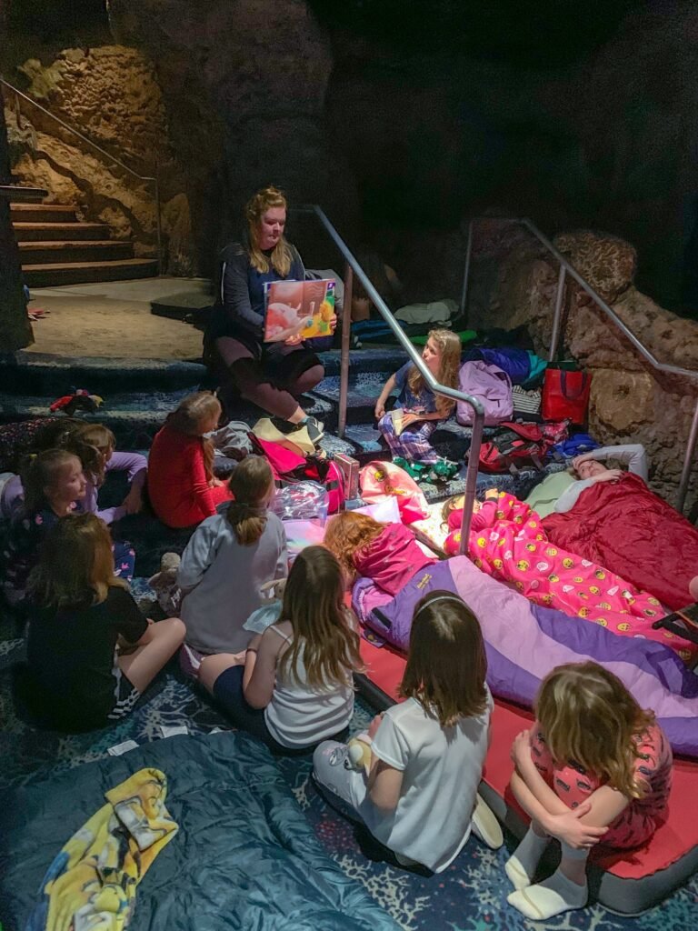 A group of children sits on sleeping bags inside a cave listening to an adult read a picture book.