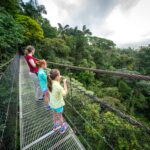Children standing on a zipline platform in a lush tropical forest under an overcast sky.