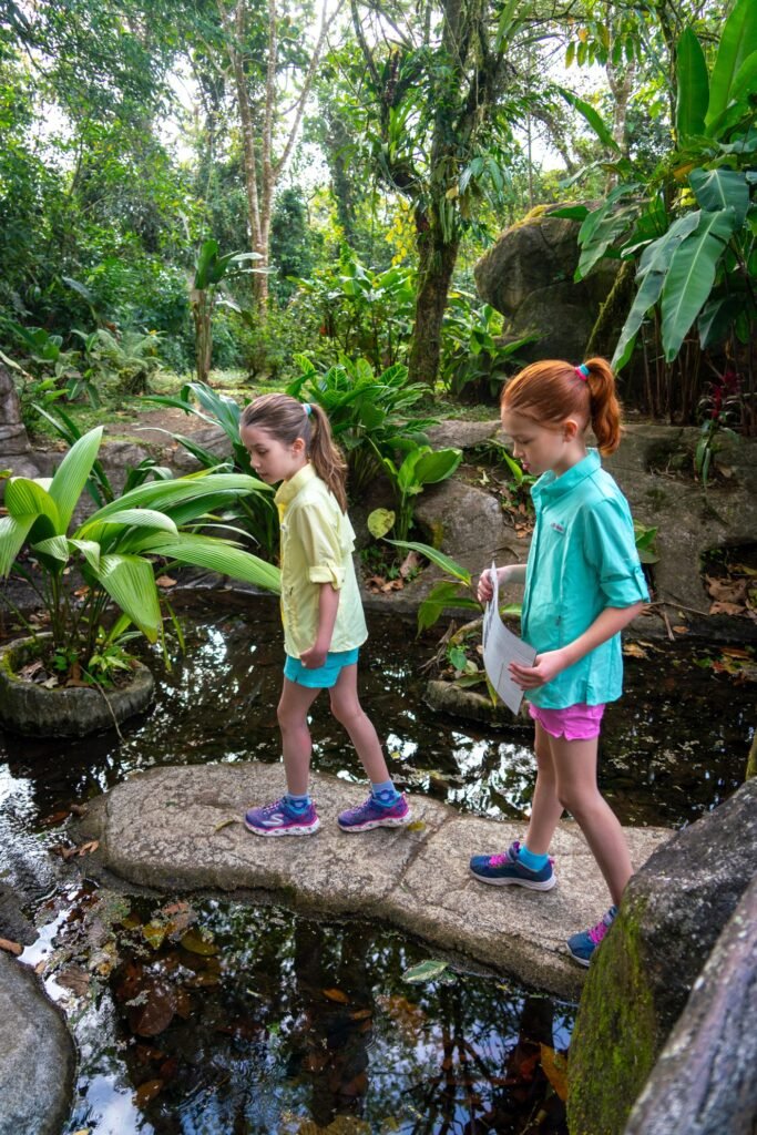 Two children crossing a stone path in a lush tropical garden. One child holds a guide map as they walk through the greenery.