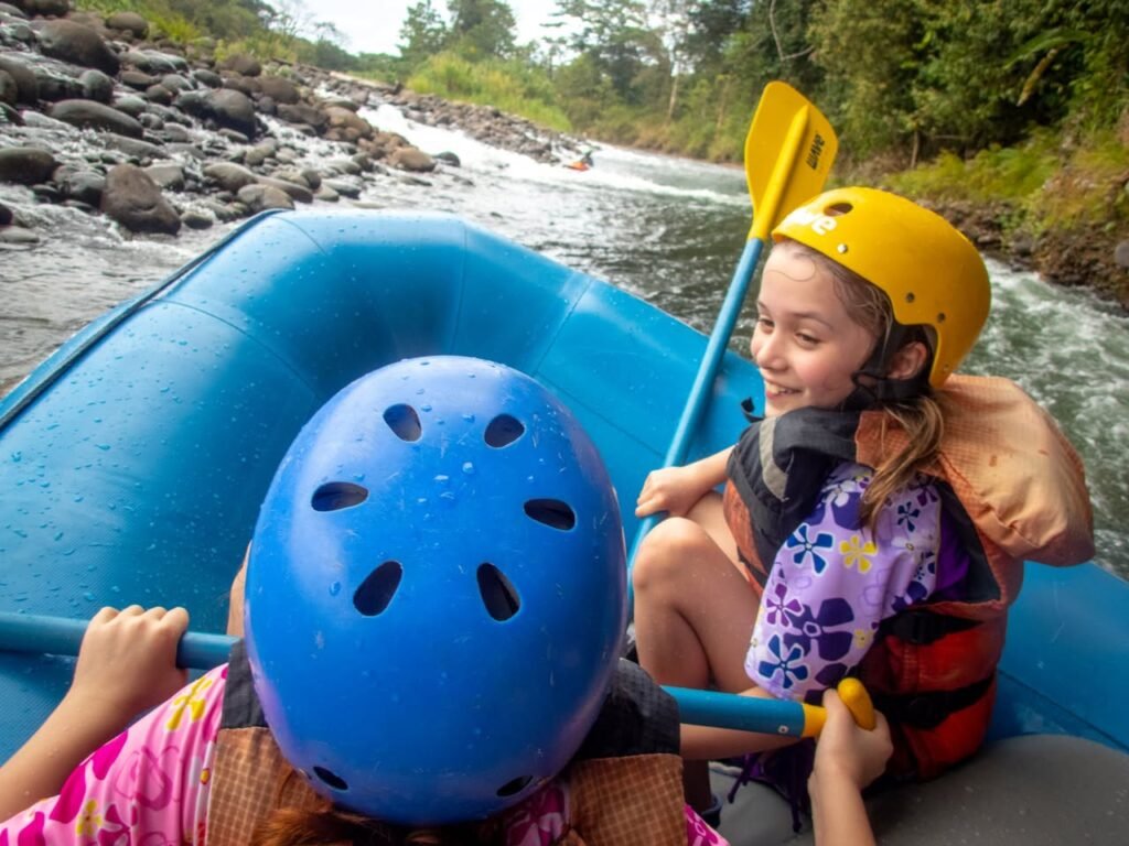 Two people in helmets paddling in a blue inner tube raft on a rocky river.