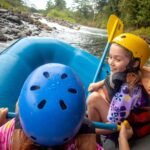 Two people in helmets paddling in a blue inner tube raft on a rocky river.