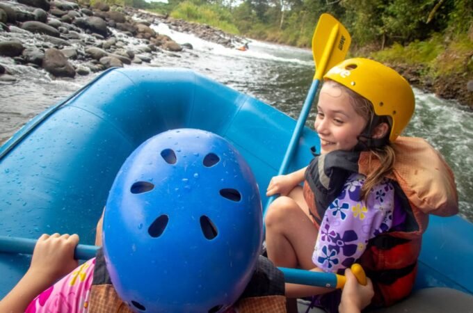 Two people in helmets paddling in a blue inner tube raft on a rocky river.
