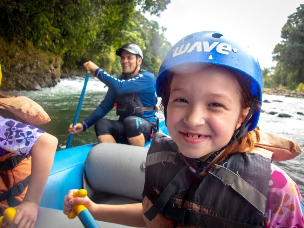 A young paddler wearing a blue rafting helmet and life jacket smiles while rafting on a river. An adult is visible paddling in the background of a lush, green outdoor setting.