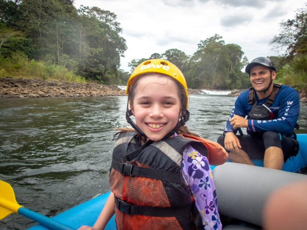 People wearing life jackets and helmets enjoy a rafting trip on a river with a forested shoreline.